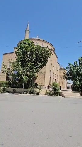 The video shows a tan brick building with a dome and a minaret under a clear blue sky. A tree with green leaves is in front of the building. The building appears to be a mosque. The ground in front of the building is paved, and there are parking spaces marked. There are also trees on the right side of the building. The video is shaky, suggesting it was filmed while someone was walking.