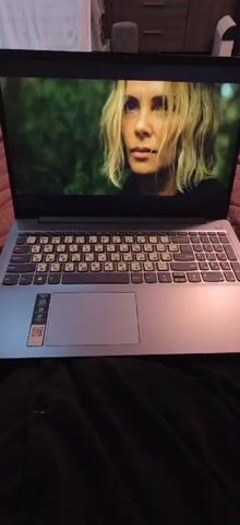 Here is a description of the video:
The video shows a laptop with a movie playing on the screen. The laptop is silver with a black keyboard that has white Russian lettering stickers on the keys. The movie appears to be a thriller or action film, showing scenes of people in a jungle-like environment and close-ups of actors with serious expressions. The laptop rests on a dark, textured surface, possibly clothing or bedding. The room is dimly lit, with the laptop screen providing most of the visible light.