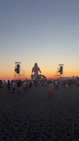 The video shows a wide shot of a beach at sunset. The sky is a gradient of deep blue at the top fading to orange and pink near the horizon. In the center of the frame, there is a large, inflatable, humanoid-shaped structure, possibly a stage prop, illuminated with orange and pink lights. On either side of this structure, tall, metal scaffolding towers are visible, each with multiple speakers mounted on them. A crowd of people is gathered on the sandy beach in front of the stage area, milling about and facing the stage. Most people are dressed in casual summer clothing. The sand is dark and appears to be wet in places, suggesting proximity to the water's edge. The overall atmosphere suggests an outdoor event, likely a music festival or concert, as the sun sets.