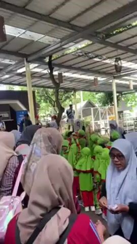The video shows a group of people, mainly women wearing hijabs, gathered under a metal roof. The environment appears to be outdoors, possibly a park or a zoo based on the glimpse of greenery and structures in the background. The women are of varying ages. Some of the women appear to be interacting with children dressed in matching green and red outfits. There seems to be an exchange of items among the women, possibly tickets or handouts. There is a person standing on a platform or elevated area in the background, seemingly giving instructions or speaking to the group. The overall atmosphere seems organized, possibly an educational outing or a guided tour.
