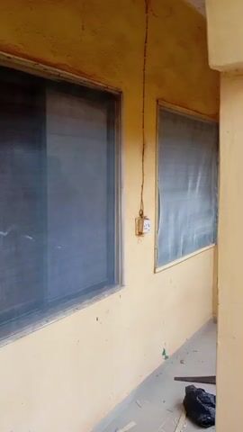 The video shows a close-up view of two windows covered with mosquito nets on the exterior wall of a building. The wall is painted a yellowish-ochre color. The window on the left is larger and appears to be a sliding window with a dark-colored mesh. The window on the right is smaller and also has a dark mesh. There is an electrical outlet with a wire running to it mounted on the wall between the two windows.

The ground in front of the building is tiled with light gray square tiles. Scattered on the ground are pieces of wood, a black plastic bag, and a handsaw with a yellow handle. The overall scene suggests an outdoor area, possibly a veranda or walkway, adjacent to the building.

The lighting indicates it is daytime, though the exact time of day is not discernible. There are no people visible in the frame. The setting appears to be in a residential or commercial area, with the mention of Rigasa, Nigeria, providing a specific location context. The presence of construction or repair materials (wood, saw) suggests that some work may be in progress or has recently occurred. There is no visible text in the video.