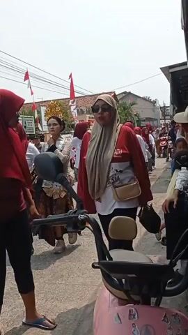 The video shows a procession or parade moving down a narrow, paved street. In the foreground, a pink scooter is partially visible. A woman wearing a red hijab and a red sweater over a white t-shirt is standing near the scooter, facing away from the camera. To her left, a young woman wearing a formal, ornate outfit with a crown is walking. Behind them, a line of people, mostly women wearing red hijabs and white t-shirts, are walking in the same direction. Some of them are carrying items. The street is lined with buildings and decorated with Indonesian flags. The weather appears to be sunny.