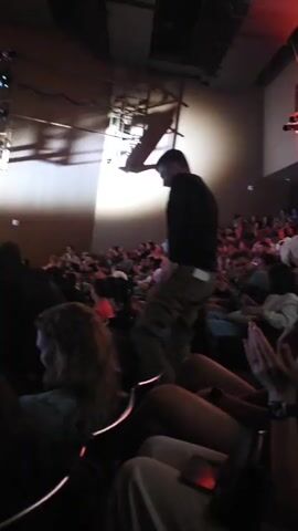 The video opens with a blurry shot of a dimly lit theater or auditorium. A large red light illuminates the upper portion of the scene, casting a warm glow. In the foreground, several people are seated, their faces partially obscured by the low light and motion blur. The camera pans slightly, revealing more of the audience. Some individuals appear to be looking towards the front, possibly at a stage or screen, while others are engaged in conversation or looking around. The overall impression is that of a crowded event taking place in a theatrical setting. The video then shifts, showing a wider view of the seated audience. The red lighting continues, creating a dramatic atmosphere. The camera moves again, capturing more close-ups of people in the audience. Some individuals are more clearly visible, showing expressions of interest or engagement. The video continues to pan and zoom, offering glimpses of various audience members and the surrounding environment. The sound of chatter and movement can be heard. The video concludes with a shot that is similar to the beginning, with a focus on the seated audience in the dimly lit space.