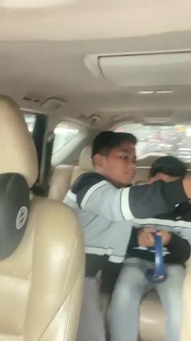 The video shows the interior of a car. In the front passenger seat, a woman wearing a purple hijab is smiling. Next to her, a baby wearing a striped shirt and a fake mustache is sitting and holding a pink bottle. In the back seat, two young men are visible. One man in a gray and white hoodie is holding a blue object, possibly a game controller. The car appears to be driving on a busy street with other vehicles and buildings in the background. The video is blurry and shaky, suggesting it was filmed on a mobile device while the car was in motion.