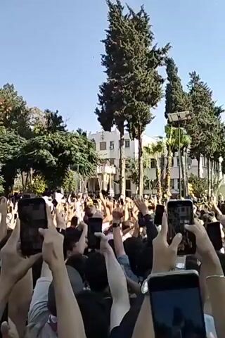 The video depicts a large crowd of people outdoors, mostly facing forward, with many holding up their cell phones, presumably recording or taking pictures. The crowd is densely packed, with arms raised high in the air. Trees are visible in the background, along with buildings, including a prominent curved glass-fronted building. The sky is clear and blue. The video is shot from within the crowd, providing a view from the perspective of someone participating in the event. The people are chanting, with the words "Allahu Akbar" audible.