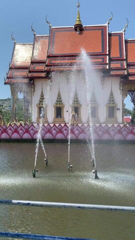 The video features an elaborate temple complex in Thailand. In the first shot, a white temple with an orange roof is seen from a distance, with water fountains spraying in the foreground. The fountains are in a body of water surrounded by pink lotus-shaped decorations. The second shot shifts to a golden statue of a mythical bird-like creature and a smaller shrine on a platform extending into the water. The final shot is a low-angle perspective looking up at an enormous statue of a smiling Buddha, situated on a lotus platform decorated with golden pillars. A small group of people, including children, are at the statue's base. The sky is clear and blue.