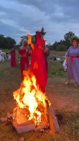 The video shows a group of people gathered around a bonfire in a grassy field at dusk. The sky is a muted blue with hints of grey clouds. In the foreground, a large bonfire burns brightly, with logs and what appears to be a cardboard box fueling the flames. Several people are visible in the middle ground and background. One woman in the foreground, on the right side of the bonfire, wears a long, blue dress and a flower crown, holding a lit object in her right hand. To her left, a person in a long, flowing red robe with a green leafy headdress dances, their arms raised. To the left of this person, a woman with blonde hair, wearing a red outfit, holds a phone and appears to be recording. Further to the left, another person in a red dress is visible. In the background, more people are scattered around, some sitting on blankets or the ground. The overall atmosphere suggests a celebratory or ritualistic gathering.