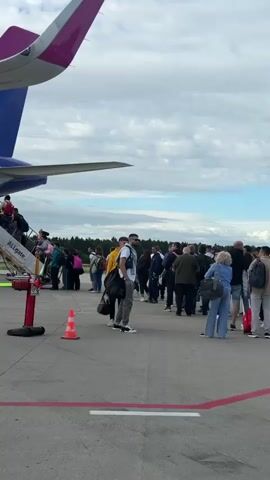 The video shows a group of people boarding a Wizz Air airplane via a mobile boarding stairs. The plane is a Wizz Air Airbus A320-271N with registration 9H-WDK. Several passengers are seen ascending the stairs with luggage. A person in a high-visibility vest is walking on the tarmac near the aircraft. Another person, facing away from the camera, is wearing a white t-shirt with graphics on the back. The scene takes place on an airport tarmac under a cloudy sky.