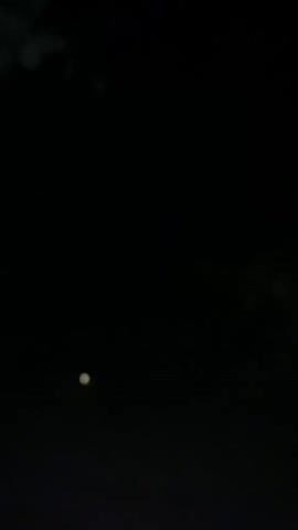 The video is extremely dark, with only a small, faint, yellow-green orb visible in the center-left of the frame. The orb appears to be stationary for the first second. Then, between the first and second second, a blurry, elongated, lighter-colored streak appears above and to the left of the orb, moving from left to right. The streak quickly disappears. The background is completely black, with the exception of what might be faint, out-of-focus foliage at the very top of the frame in the first second, and a slightly less dark, dark blue-grey hazy area at the top of the frame from the second second onwards. There are no other discernible objects or figures. The overall impression is of a nighttime scene with very low light conditions.
