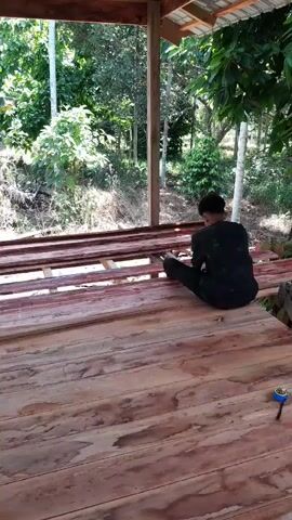 The video shows a man working on a wooden structure under a covered shelter. The shelter has a wooden frame and a roof, with an open view of a lush, green environment filled with trees and banana plants. The man is sitting on the wooden planks, wearing a dark, patterned shirt and dark pants. He appears to be hammering or adjusting something on the wooden structure. A small green bucket and a red cloth are visible nearby. The sound of hammering can be heard throughout the video.