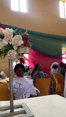 The video shows an indoor gathering of people seated at tables, likely a celebration or event. In the foreground, a woman in a white lace outfit is seated, looking towards the camera. Behind her, other women are also seated, some looking forward, others to the side. The room is decorated with draped fabric in pink and green hues, and a floral arrangement is visible on the left side of the frame. Sunlight streams in from windows at the top of the image. The overall atmosphere appears to be formal and celebratory.