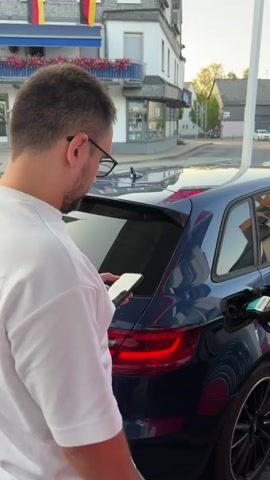 A man in a white t-shirt with "ESSENTIALS" printed on the back is standing next to a dark blue Audi A3 Sportback at a gas station. He is holding a smartphone in his left hand and appears to be looking at it. The car's fuel door is open, and a gas pump nozzle is inserted into it. The car has a German license plate that reads "AO 827".

The scene is set in Sohren, Germany, as indicated by the German flags visible on the building in the background. The building also features a balcony with flowers and signage. The gas station itself has a white pole with a red and white sign at the top and a yellow diamond-shaped road sign to the right, suggesting it is a priority road. Other buildings and trees are visible in the distance. The lighting suggests it is late afternoon or early evening, with a warm, soft glow. The weather appears to be clear and pleasant. The overall mood is one of everyday routine, with the man engaged with his phone while refueling his car.