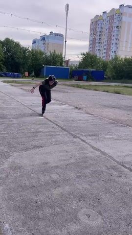 In this video, a person is rollerblading in an outdoor urban environment. The subject is wearing a helmet, a dark t-shirt with a camouflage pattern, and dark pants with a red stripe down the side. They are in a crouched position, appearing to be maneuvering or practicing a skating technique. The rollerblades are black and appear to be aggressive or freestyle skates.

The scene is an open, paved area, possibly a disused parking lot or a skate park. The ground is concrete with visible cracks and markings. In the background, there are trees, some buildings, and a utility pole with lights. The buildings appear to be residential, with multiple stories and windows. There's also a glimpse of a blue structure and some scaffolding or metal framing. The sky is partly cloudy, suggesting it is daytime, likely afternoon given the lighting. The overall impression is a casual, everyday activity taking place in a typical Russian urban setting. There is no visible text in the video.