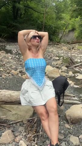 A woman with blonde hair and sunglasses is posing for a video outdoors. She is sitting on a log near a rocky stream with trees in the background. She is wearing a blue polka-dot bandana as a top and a light-colored skirt. Her arms are raised behind her head.