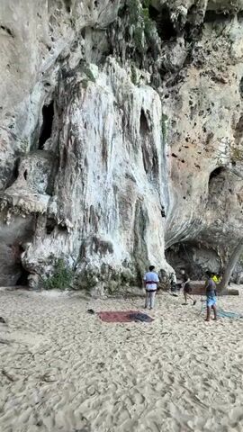 A video shows a rock climber ascending a large, textured limestone cliff face with a cave opening visible. The climber is a dark silhouette against the lighter rock. Below the cliff, on a sandy beach, several people are gathered. One person, wearing a white shirt and beige pants, is standing near a red climbing mat. Another person, wearing a black shirt and shorts, stands beside them. A third person in the foreground, wearing a blue patterned shirt and blue shorts, is facing away from the camera. Further down the beach, more people are visible near the water. A tree with green foliage grows from the cliff face to the right of the climber. The scene appears to be a popular spot for outdoor activities, likely rock climbing and beach recreation.