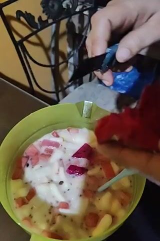 A person is cutting a piece of red dragon fruit and adding it to a bowl containing a mixture of fruits and a creamy substance. The bowl is light green. The piece of dragon fruit is bright pink on the outside and dark red inside, with black seeds. The person uses a black handled knife. A small light blue plastic spoon is in the bowl with the mixed fruits. The table is a dark, reflective surface. A blue plastic bag is visible in the background.