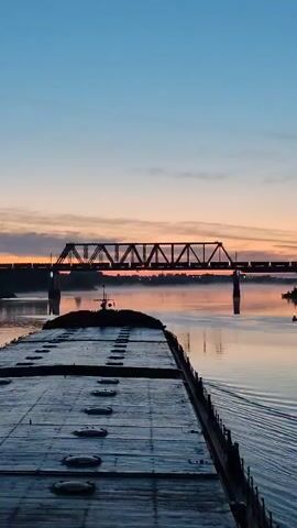 The video shows the perspective from a barge moving down a calm river at sunset. The sky is blue fading into orange near the horizon. A metal railroad bridge spans the river in the distance, with a train visible on it. The barge has a flat, metallic surface with evenly spaced circular hatches. Wake patterns trail behind the barge, indicating its movement. The overall environment is serene, captured during twilight hours.