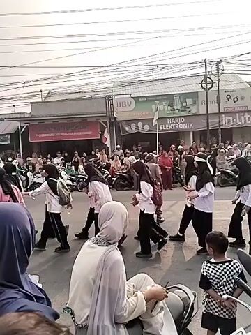 A group of people, predominantly young women in dark headscarves and light-colored tops, are walking across a street. Some carry backpacks. They are walking in a procession, moving from left to right in the frame. In the background, there are buildings with signs, including one that says "HARJO ELEKTRONIK." Several adults, some on motorcycles, are watching the procession. A young boy in a checkered shirt is also visible. The environment appears to be a commercial street with overhead power lines. The lighting suggests it is daytime. The overall scene depicts an organized movement of people through a public area, likely a parade or a school-related activity.