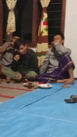 This video shows a group of men sitting on a blue tarp on the floor. They are in a room with white walls and a window. Some of the men are wearing traditional Islamic clothing. One man is writing in a notebook, while others appear to be talking or eating. There are plates of food and drinks on the tarp. The lighting is dim, suggesting it is nighttime.