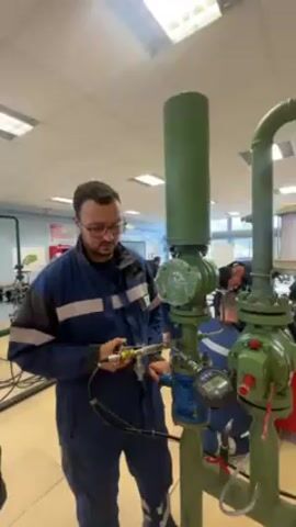 A man in a blue jumpsuit stands in a room with multiple people in the background. He’s holding a silver-colored tool and pressing a button on it, which makes a hissing sound. The man is standing in front of pipes that are painted green, which appear to be part of an industrial setup. There’s also a digital display attached to the pipe. He looks at the device in his hand, then looks directly at the camera and speaks. Another voice can be heard telling him to make the device “sing,” and he responds.