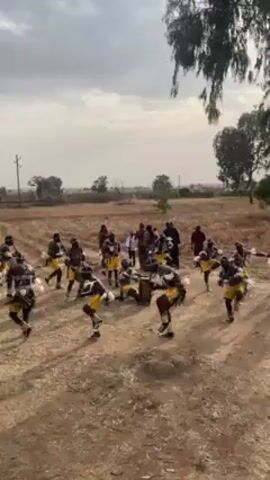In a medium-shot, eye-level video, a group of people performs a traditional dance in a dry, rural setting. The dancers, numbering around twenty, are dressed in black headgear and tops, yellow shorts, and adornments of white feathers or fur around their waists and calves. They are arranged in a circular formation, moving in rhythm with each other. In the center of the circle, some individuals are drumming, providing the beat for the dance. Behind the dancers, several people stand watching, dressed in a variety of casual clothing. The ground is dirt, with sparse patches of dry grass and stubble. In the background, there's an open field dotted with a few trees and utility poles. A large tree with drooping branches frames the upper right corner of the shot. The sky is overcast.