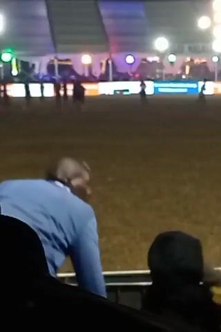 The video depicts a nighttime outdoor scene, likely at an event, based on the visible crowd and stage lighting in the background. The primary focus is on several people in the foreground. A man in a blue jacket and brown pants initially has his back to the camera. Then, a person wearing a grey hooded sweatshirt approaches the man in the blue jacket. A brief interaction appears to occur between them. The environment consists of a grassy field with a stage or elevated structure illuminated with bright lights and video screens in the distance.