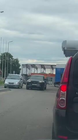 The video shows a view from inside a car at a gas station. There are several cars lined up at the pumps. The weather appears overcast. The cars have Russian license plates. One car in the foreground is a black Lada. Another car in the middle ground is a light blue SUV. Further back, a black sedan is visible. The gas station canopy and pumps are also visible.