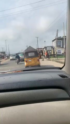 A daytime dashcam perspective captures a bustling street scene with moving vehicles and pedestrians. The camera is positioned inside a car, looking forward through the windshield. A yellow three-wheeled auto-rickshaw is prominently visible in the foreground, followed by another in the midground. A blue auto-rickshaw is seen on the right side of the road, and another yellow auto-rickshaw appears on the far right. A white car and a silver car are visible in the distance, interspersed with motorcycles. A man in a red shirt and black pants is walking on the right side of the road near a blue auto-rickshaw. Another man in a black shirt and beige pants is standing on the far right. Utility poles and power lines crisscross the sky, with residential buildings and shops lining the roadside. The sky is partly cloudy, and the overall lighting suggests a bright day.