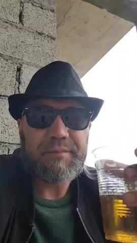 A man in a black fedora and sunglasses, with a grey beard, takes a sip of a golden beverage from a clear plastic cup. He is wearing a dark jacket over a green sweater. The background suggests an outdoor setting with a textured concrete wall to his left and a concrete overhang above. The lighting indicates it might be daytime, possibly overcast. The man's expression is neutral, focused on drinking. The scene appears to be casual and relaxed.