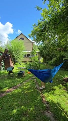A person in light-colored pants and socks is getting into a blue hammock strung between two trees in a backyard. The hammock is positioned in front of a two-story yellow house with a green roof and white trim. The house has several windows and a large arched window on the second floor. The yard is covered in green grass and surrounded by lush green trees and bushes. To the left of the hammock, there is a small grill, a white table with some items on it, and a wooden structure. The sky is blue with some white clouds. The sun is shining, casting shadows on the grass.
