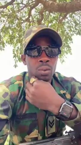 A man wearing a camouflage uniform, cap, and dark sunglasses is looking at the camera with a serious expression. He has a gold ring on his right hand and a digital watch on his left. He has a goatee and is touching his chin with his hand. He appears to be sitting under a tree with light green leaves, with a clear sky visible in the background. A rifle is visible near the bottom of the frame.