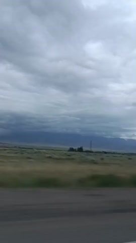 The video shows a view from inside a vehicle moving along a paved road. The road has a white line on the right side. To the left of the road is a vast, flat field of dry grass and some scattered trees. In the distance, there are low mountains under a cloudy sky. The clouds are heavy and gray, with some patches of blue sky visible. The overall impression is that of a rural landscape, possibly during the daytime or dusk, with signs of an approaching storm.