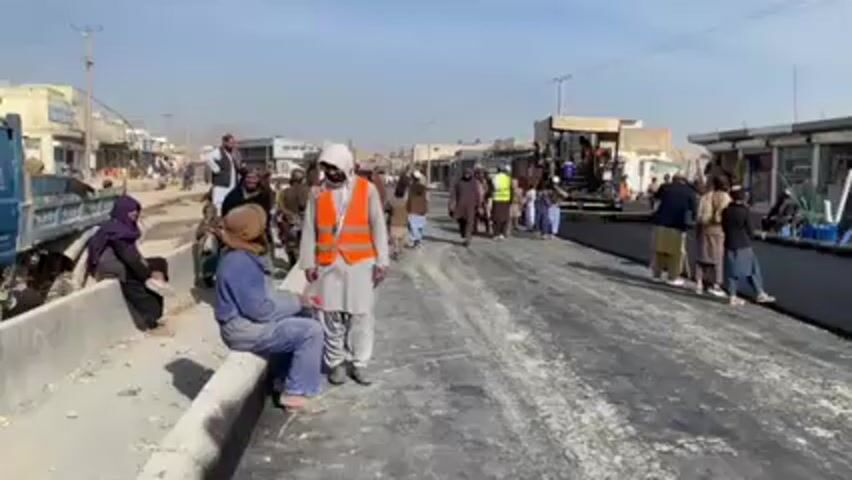 The video shows a street scene in what appears to be a Middle Eastern or South Asian town, where road construction is taking place. A large paving machine is visible in the middle of the road, laying down a fresh layer of asphalt. Several people are gathered around the construction site. Some are watching the paving process, while others appear to be workers involved in the project. There are both men and women present, some wearing traditional clothing. The buildings lining the street appear to be shops or commercial establishments. The overall atmosphere suggests a community observing and participating in infrastructure development. The sky is clear, indicating a sunny day. There are utility poles and lines visible in the background.