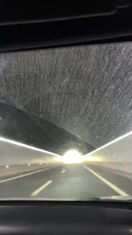 The video shows a view from inside a car as it travels through a tunnel. The car's windshield is visible in the foreground and has a dirty, streaky appearance. The tunnel walls are lined with lights, and the road ahead is dark, leading to a bright exit. The perspective is from the driver's seat, looking forward through the windshield.