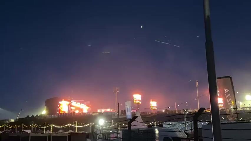The video shows a long shot of an outdoor event at dusk, with several brightly lit stages and screens in the distance. The sky is dark blue, transitioning to a lighter shade towards the horizon. The stages are glowing with orange and red lights, with some smoke or mist visible around them. In the foreground, there are string lights, fences, and various structures. A black and white banner with the word "ASSA" partially visible on it is seen on the right side of the frame. The overall impression is that of a large music festival or concert.