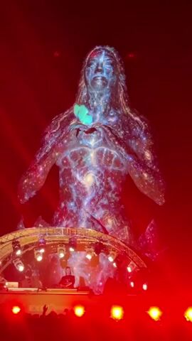 The video shows a large projection of a female figure on a stage. The figure is adorned with colorful, swirling patterns that resemble galaxies and nebulae, and at times with flowers. In the foreground, a DJ booth is visible with a person operating equipment. The stage is lit with bright, dynamic lighting, predominantly red, which shifts and changes throughout the video. A crowd of people can be seen in silhouette at the bottom of the frame, watching the performance. The projection changes its appearance, cycling through different visual themes.