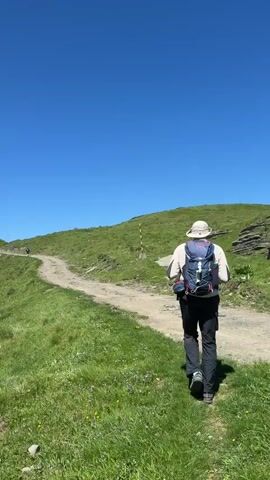 The video shows a scenic landscape on a clear, sunny day. The camera pans across the landscape starting from a dirt path winding through a grassy hillside. A person is visible on the path in the distance. The camera pans to reveal rolling green hills and mountains in the background, some of which are snow-capped. The camera continues to pan revealing more grassy slopes and a view of a distant body of water in the horizon.