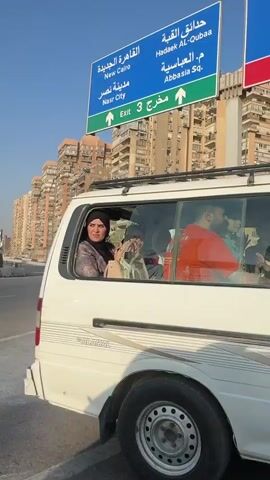 The video captures an urban scene from an elevated road or highway, likely in Shubra al Khayma, Egypt, during a sunny day with a clear blue sky.

The primary subject is a white passenger van, identifiable as a "KING LONG" brand vehicle, carrying several occupants. Inside the van, a driver is visible, along with passengers including a woman wearing a hijab, a man in a red shirt, and children. One man in a light green shirt is seen smiling and waving at the camera.

Overhead road signs, prominent and blue with white text, provide directional information in both Arabic and English. These signs indicate routes to "Khalafawy Sq.," "Shubra St." (Exit 2), "New Cairo," "Hadaek Al-Qubaa," "Abbassia Sq.," and "Nasr City" (Exit 3).

In the background, numerous tall, multi-story residential buildings with multiple balconies are visible, typical of a dense urban environment. The buildings appear to be made of light-colored concrete or stucco. The van itself shows some minor cosmetic damage on its front bumper and displays the Arabic numbers "٢٤١٢" (2412) near its headlight. The overall scene depicts a busy road in an Egyptian city.