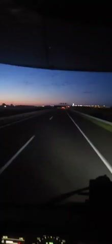 A first-person perspective video shows a road at dusk. The sky transitions from a deep blue at the top to a lighter orange and pink hue near the horizon. Dark clouds are scattered across the sky. The road ahead is dark and empty, with white lines marking the lanes. On the right side of the road, a yellow guardrail is visible. A side-view mirror of a vehicle is visible on the right side of the frame, reflecting some lights. There are no people visible in the video. The overall environment appears to be a highway or a similar road in a rural or undeveloped area.