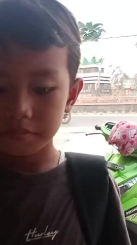A close-up of a child's face is visible from the side, with their head turned downwards. In the background, a bright green motorcycle is parked, draped with a pink cloth decorated with white hearts and faces. Further in the distance, a large pineapple monument stands prominently at an intersection, surrounded by traffic lights and flags. The overall scene suggests an outdoor setting, possibly a roadside or a public square.