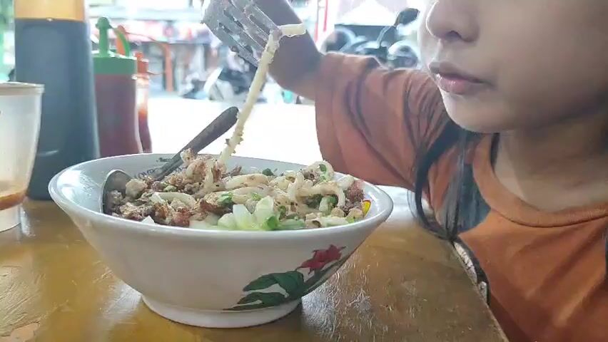 A close-up shot shows a young girl eating noodles from a bowl. She is using a fork to lift a portion of the noodles to her mouth. The bowl of noodles contains chopped green onions and other ingredients. In the background, a bottle of soy sauce and a bottle of ketchup can be seen. The girl is wearing an orange shirt. The scene appears to be in a restaurant setting.