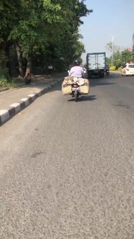 The video shows a point-of-view shot from a moving vehicle on a road. The primary focus is a person riding a motorcycle ahead, carrying a large load strapped to the back. The rider is wearing a helmet and a light-colored shirt. The motorcycle is loaded with items wrapped in what appears to be light-colored fabric or plastic. The road is paved and appears to be a two-lane road with a sidewalk and trees lining one side. Other vehicles, including a large truck and a white car, are visible on the road in the distance. The environment appears to be an urban or suburban setting with buildings and vegetation visible. The sky is clear and the lighting suggests it is daytime. The camera is in motion, following the motorcycle.