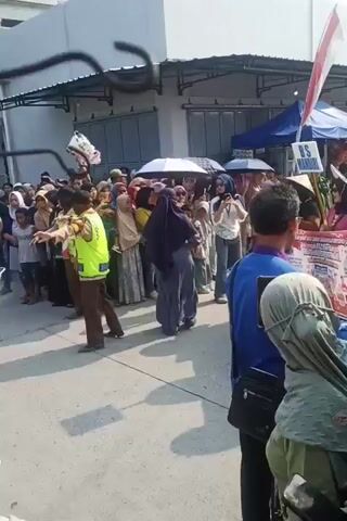 A street scene with many people gathered. Most of the people are facing the same direction, suggesting they are watching something. There are several tents and stalls visible in the background, indicating some sort of event or market. Some individuals are holding umbrellas, possibly for shade or protection from the sun. Flags and banners are present, including an Indonesian flag. People are dressed in a variety of clothing, some in traditional attire. There are a few individuals holding up their phones, likely to record or photograph the event. The overall atmosphere appears to be lively and crowded.