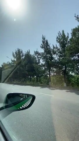 The video is filmed from inside a vehicle, looking out the side window and reflecting the side mirror. The side mirror shows a checkered pattern on its edge and a person inside the car wearing headphones and possibly sunglasses. Outside the car, a road with cracks and some debris is visible. On the left side of the road, there are several tall trees, a street lamp, and some vegetation. The sky is bright and sunny, with a visible sun flare in the upper left corner. In the distance, there are more trees and some structures, including a white truck. The car appears to be moving.