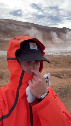 A short video captures a close-up selfie of a young person, likely male, in an outdoor, rugged landscape identified as Reykjahlíð, Iceland. The subject is wearing a vibrant orange hooded jacket with black accents and a black baseball cap featuring a white "Carhartt" logo. They hold a white smartphone in a clear case in their left hand, seemingly recording themselves. Their right index finger is pressed against their nose, slightly pulling down the jacket's collar. A metallic watch is visible on their left wrist.

The background reveals a barren, earthy terrain with brown hills and mountains under an overcast sky. Several plumes of steam or smoke rise from various points in the distance, indicative of geothermal activity common in Iceland. The overall impression is of a cool, possibly windy, day in a unique geological setting. The person's direct gaze at the camera suggests engagement, and the nose-touching gesture could be a playful reaction or a response to the sulfurous smell often present in geothermal areas.