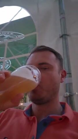 A man with short brown hair and stubble is seen from the chest up, wearing a coral colored polo shirt with a blue collar. He is holding a glass of beer to his lips and drinking it. The background is out of focus, but appears to be an outdoor setting with a large white tent or awning overhead, and a green umbrella visible in the distance. A metal pole is also visible in the background, to the right of the man's head.