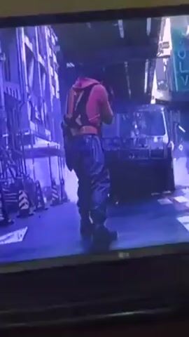 The video shows a man in a red hoodie and blue pants walking towards a black sports car parked on a street with blue-tinged lighting. The car's taillights are visible, and there's graffiti and urban elements in the background. Later, the scene shifts to a man in a clear raincoat handling items, possibly packages or boxes, covered with colorful patterns. The video is recorded from a television screen, with the LG logo visible at the bottom. The image quality is somewhat grainy due to the recording. An Arabic caption is displayed at the bottom of the screen.