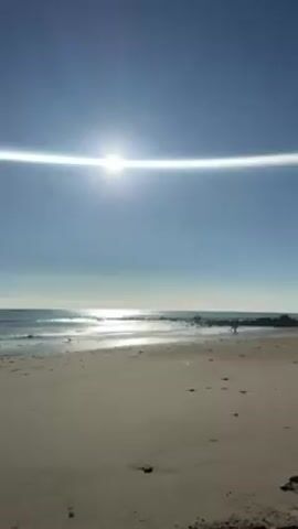 The video shows a beach scene on a sunny day. The sky is clear and blue, with the sun shining brightly and creating a light flare effect. The sand on the beach is light-colored and appears damp near the water. Small waves are visible in the ocean. In the distance, a rock jetty extends into the water, and two people are walking on it.