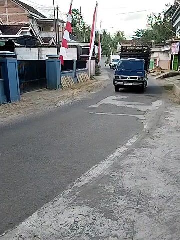 A black Mitsubishi pickup truck, loaded with coconuts, drives down a paved road. The truck is positioned in the center of the road. The truck is moving from left to right in the frame. The truck is heading towards a slight bend in the road, which is lined with trees and vegetation. The sky is overcast and the lighting suggests it is daytime. Buildings and houses are visible on the left side of the road. There are flags on poles in front of one of the buildings. The truck is driving away from the camera.