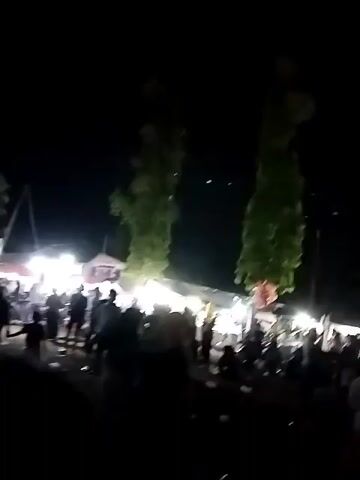 The video shows a nighttime outdoor gathering with many people. Bright lights illuminate tents and stalls, suggesting a market or festival. People are milling about, and some appear to be dancing or watching a performance. The atmosphere seems lively.